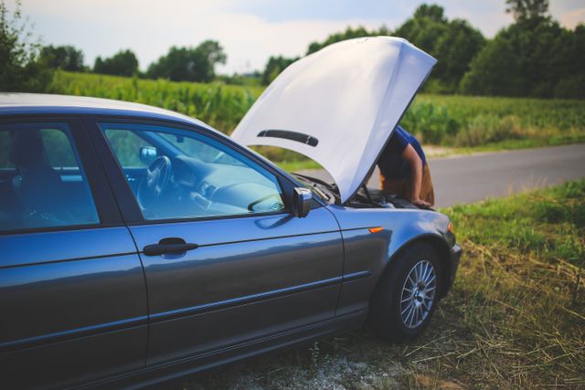 Sivi automobil sa podignutom haubom i osobom koja gleda u motor pored puta uz šumu